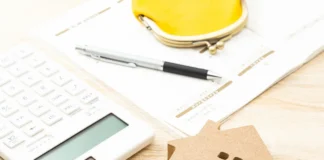 Kalkulator, notes, żółty portfel i drewniany domek ułożone na biurku jako elementy planowania budżetu domowego